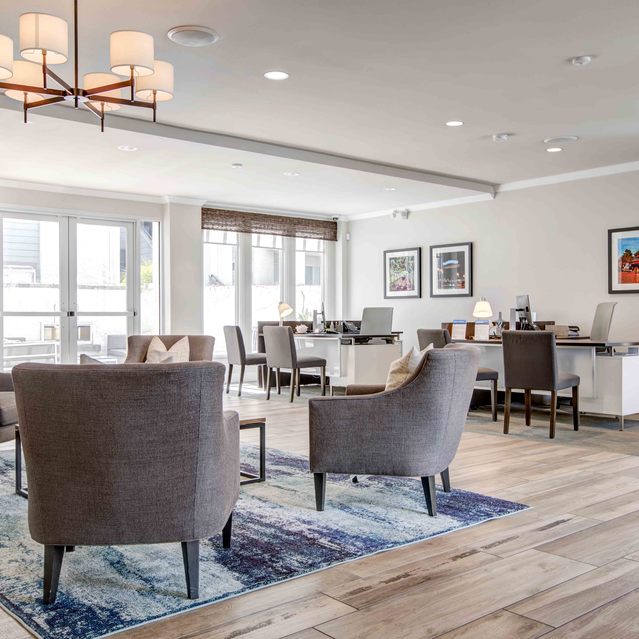 Stoneridge – Elegant community lounge with gray armchairs arranged on a blue patterned rug, adorned by a modern chandelier